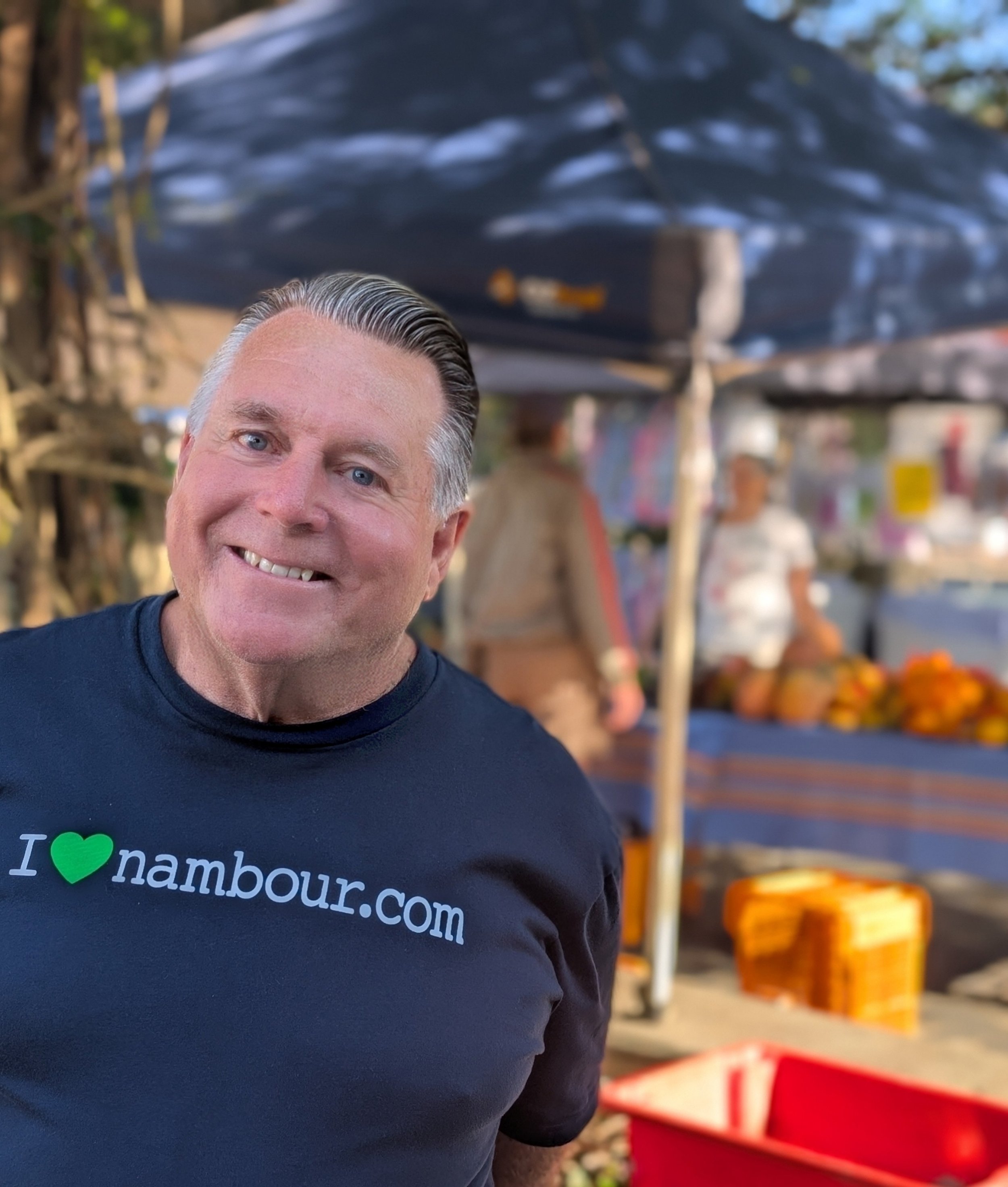 Close-up of a smiling man with gray hair wearing a dark blue shirt with the text 'I ❤️nambour.com'. In the background, there are shelves with various products, including orange and yellow items, and what appears to be a market or store setting.
