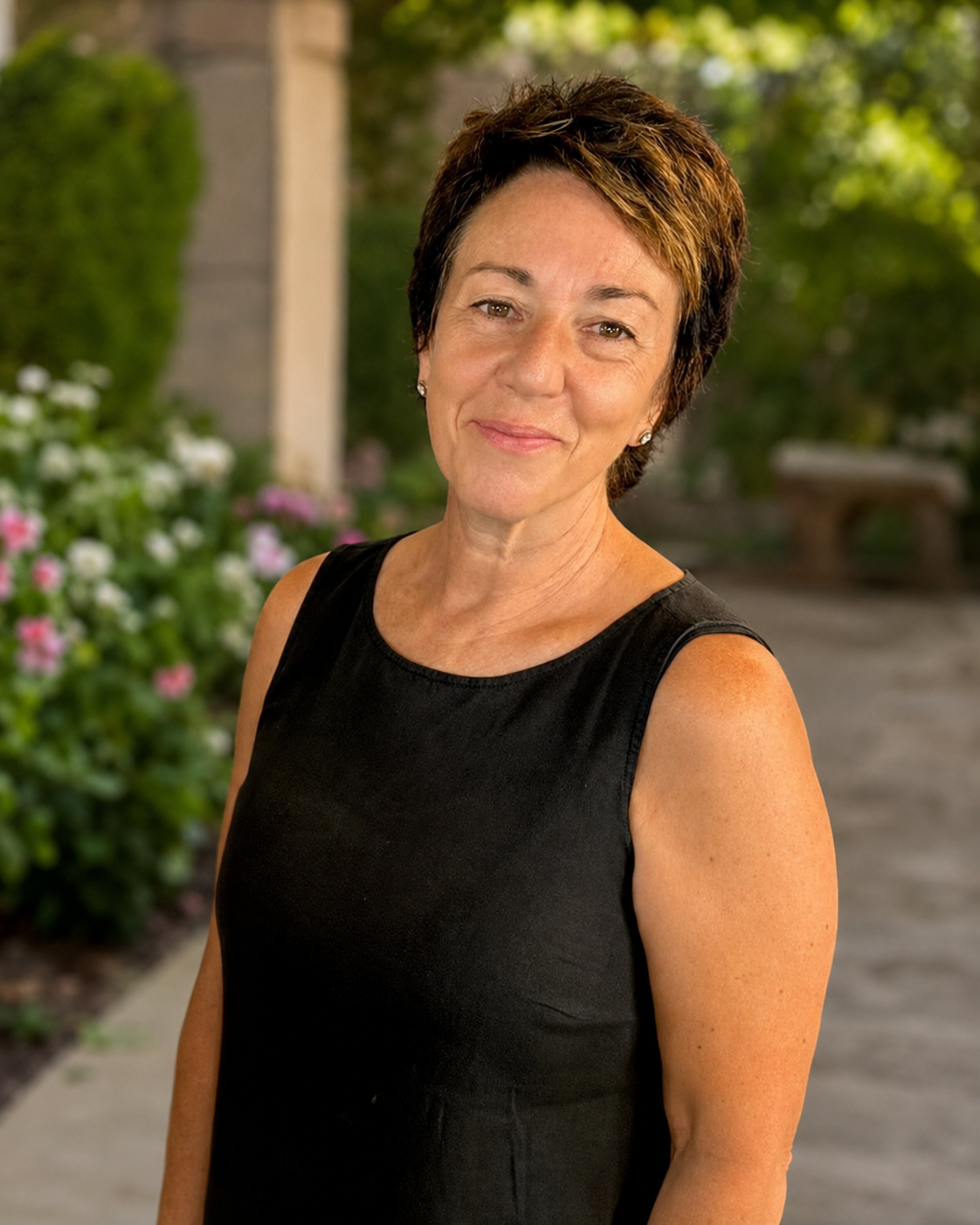 A middle-aged woman with short brown hair styled with highlights, wearing a black sleeveless top, standing outdoors on a sidewalk with blurred garden and greenery background.