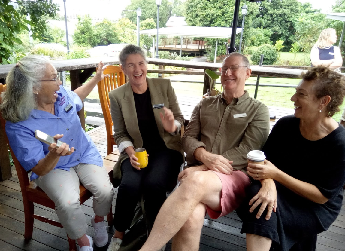Four people sitting on a wooden patio, laughing and enjoying a moment together, with green trees and a gazebo in the background.