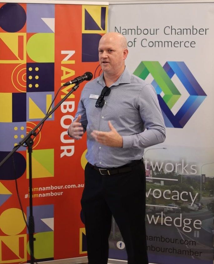 A man speaking at a microphone during a Nambour Chamber of Commerce event, with colorful banners in the background.