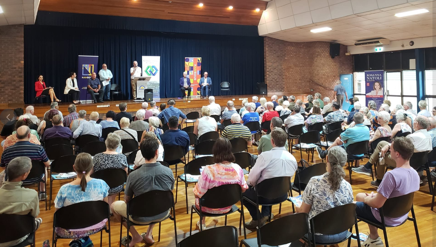Audience attending a panel discussion on a stage in a large auditorium with seated officials and speakers, and banners in the background.