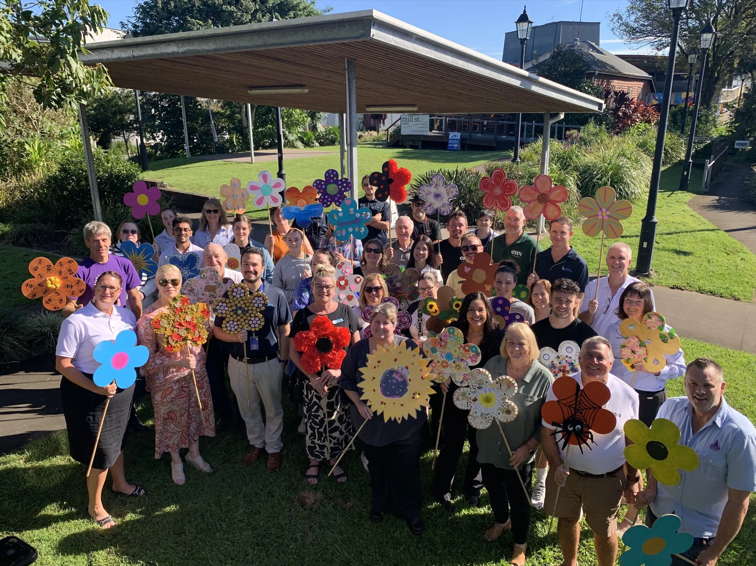 Group of people outdoors holding colorful paper flowers and decorations, smiling at the camera during daytime.