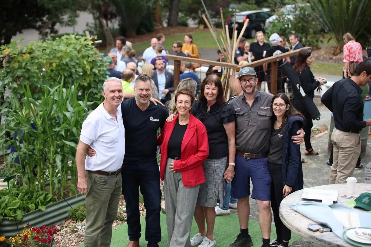 Group of six people smiling and posing together at an outdoor gathering, with more people in the background, surrounded by greenery and a wooden windmill decoration.