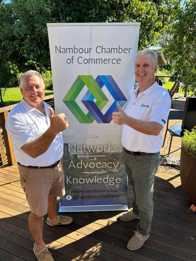 Two smiling men giving thumbs up next to a Nambour Chamber of Commerce banner outdoors.