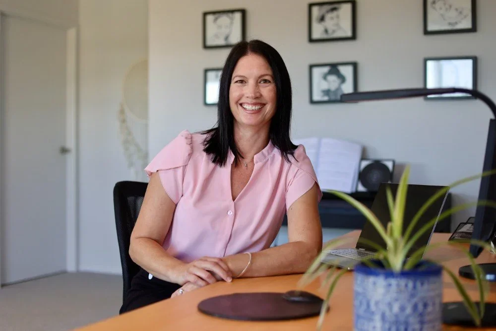 Smiling woman with shoulder-length dark hair, wearing a pink blouse, sitting at a desk in an office with framed artwork on the wall.