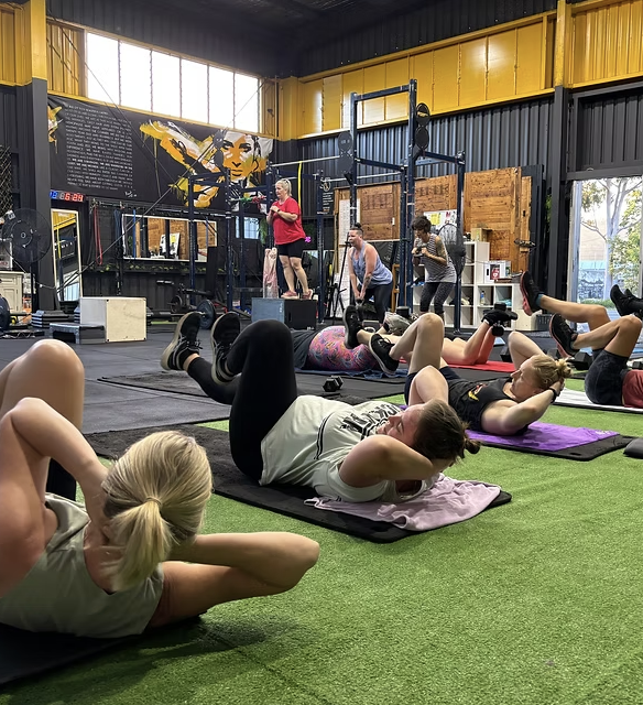 Group fitness class in a gym with women lying on mats doing abdominal exercises, instructor standing on a platform leading the class, inside a brightly lit gym with black and yellow walls, and large windows.
