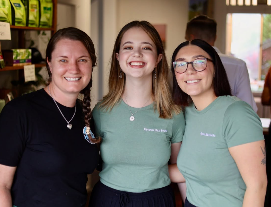Three smiling women standing inside a store, two wearing green t-shirts and one wearing a black shirt, with shelves and other people in the background.