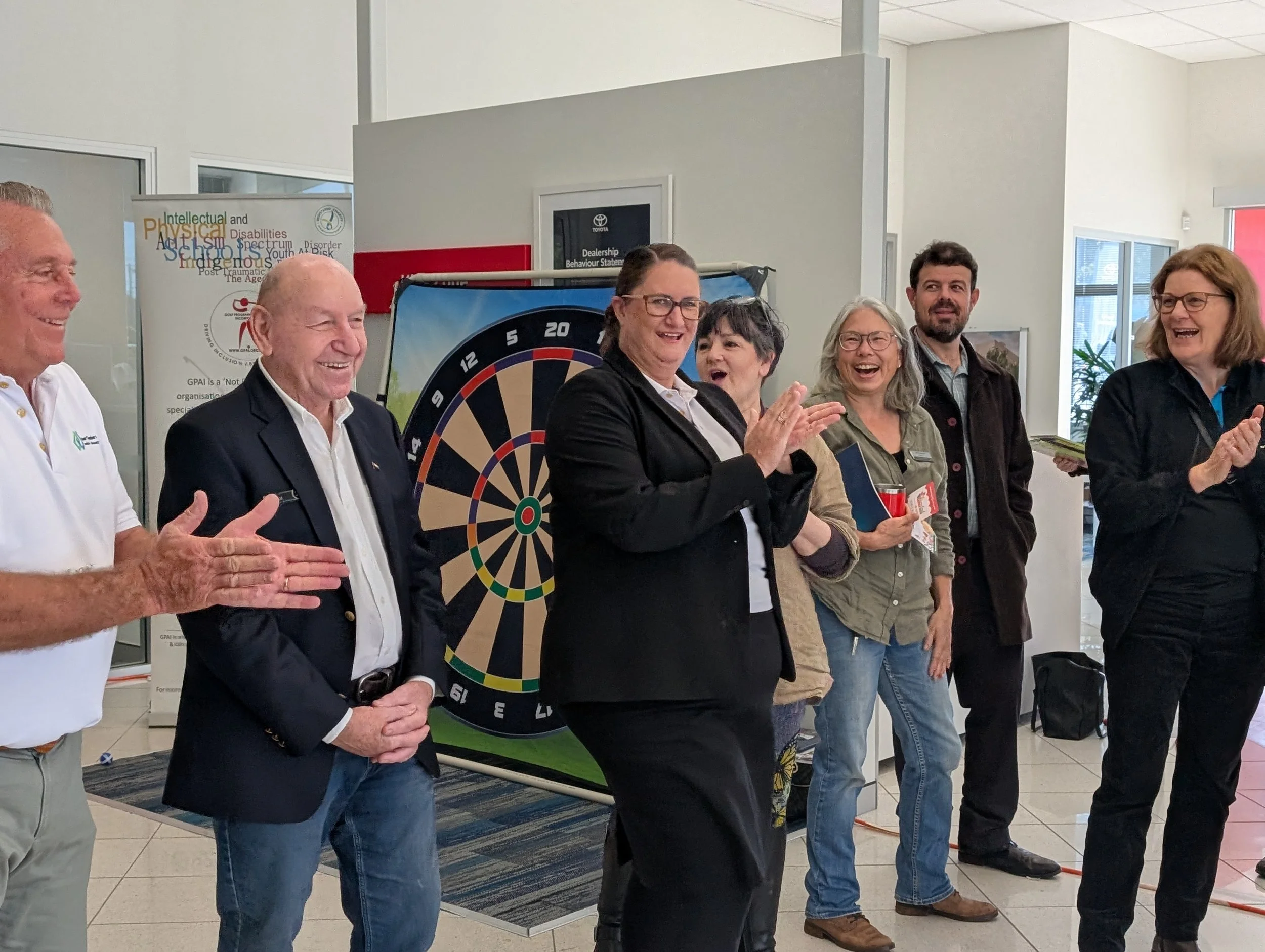 Group of people standing in a line, clapping and smiling, in an indoor setting with a dartboard and banners in the background.