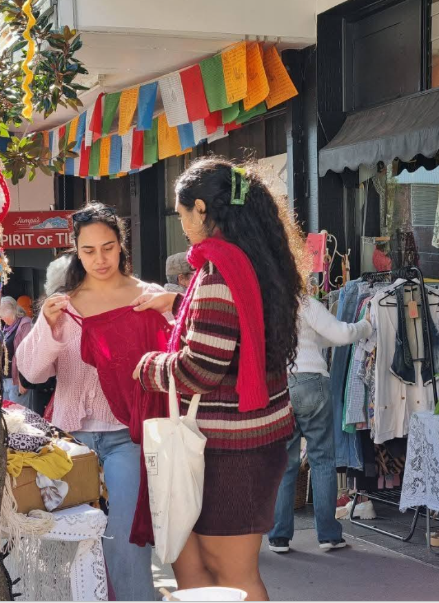 Two women shopping at an outdoor market. One woman with long dark hair wearing a red and beige striped sweater and a red scarf holds a red dress, while the other woman with long dark hair in a pink sweater looks at the dress. Behind them, there are c