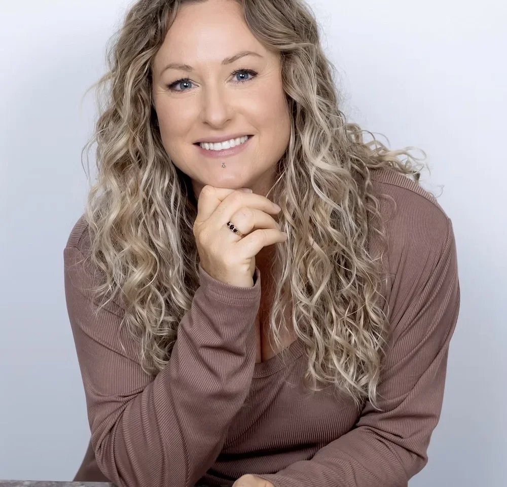 A woman with curly blonde hair, wearing a brown long-sleeved shirt, smiling and resting her chin on her hand with a ring and lip piercing, against a plain white background.