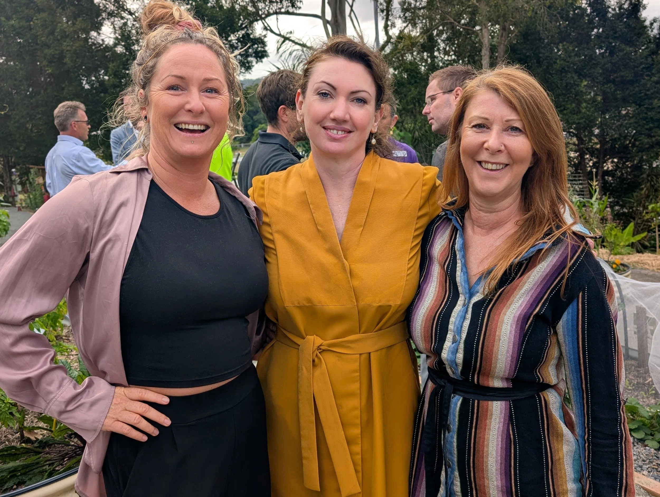 Three women smiling and posing for a photo outdoors, with trees and other people in the background.