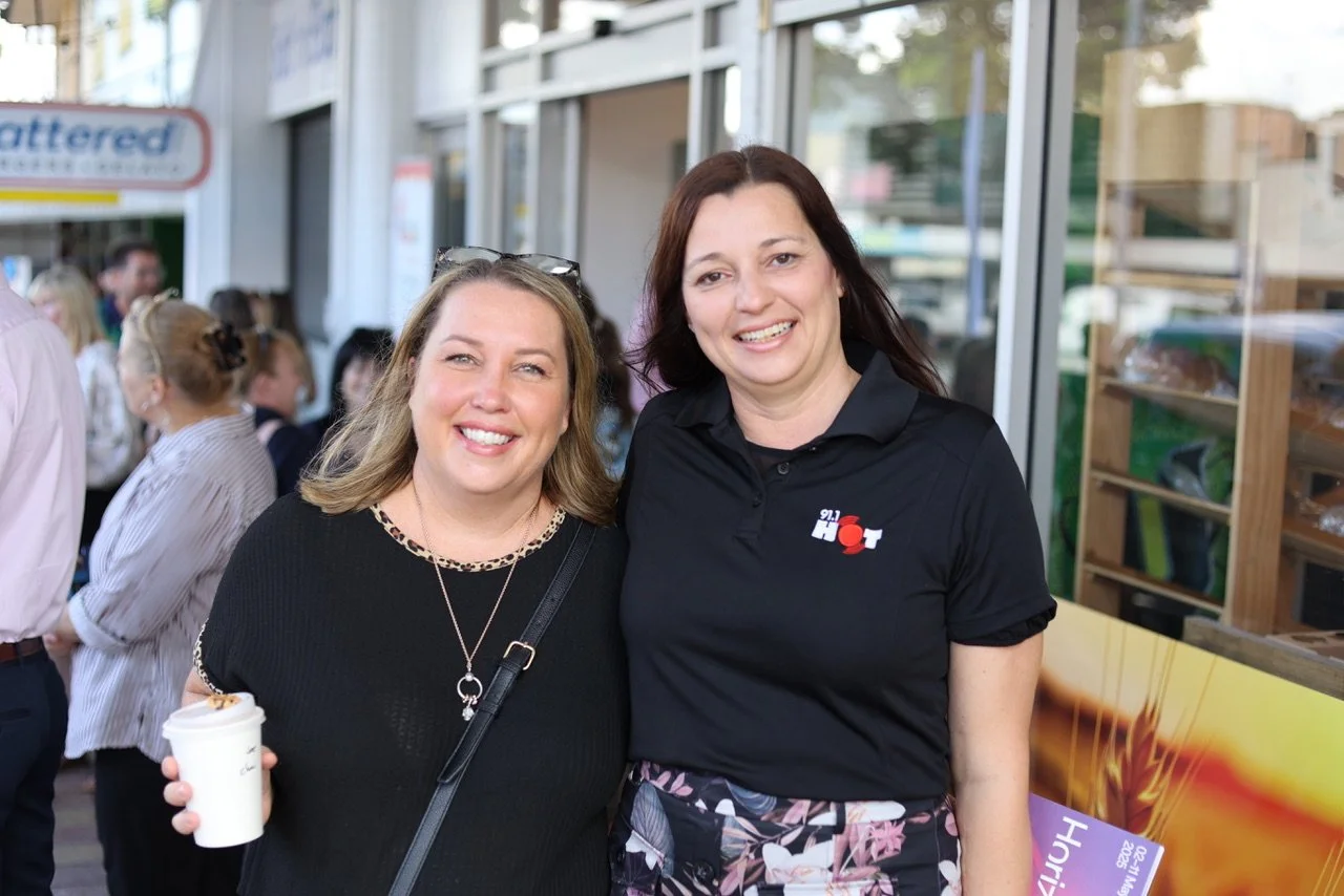 Two women smiling and posing for a photo outside a building, with several people in the background.