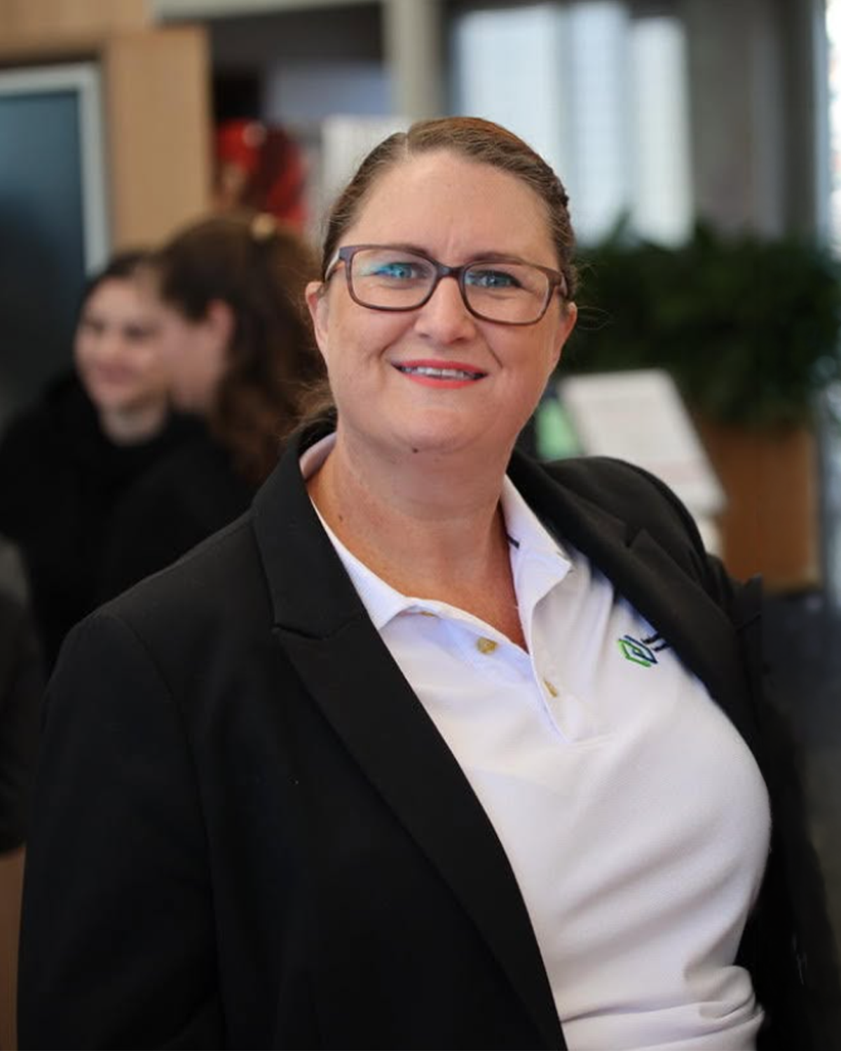 A woman with glasses and brown hair styled back, smiling, wearing a black blazer and white polo shirt, indoors with blurred background of another woman and office setting.