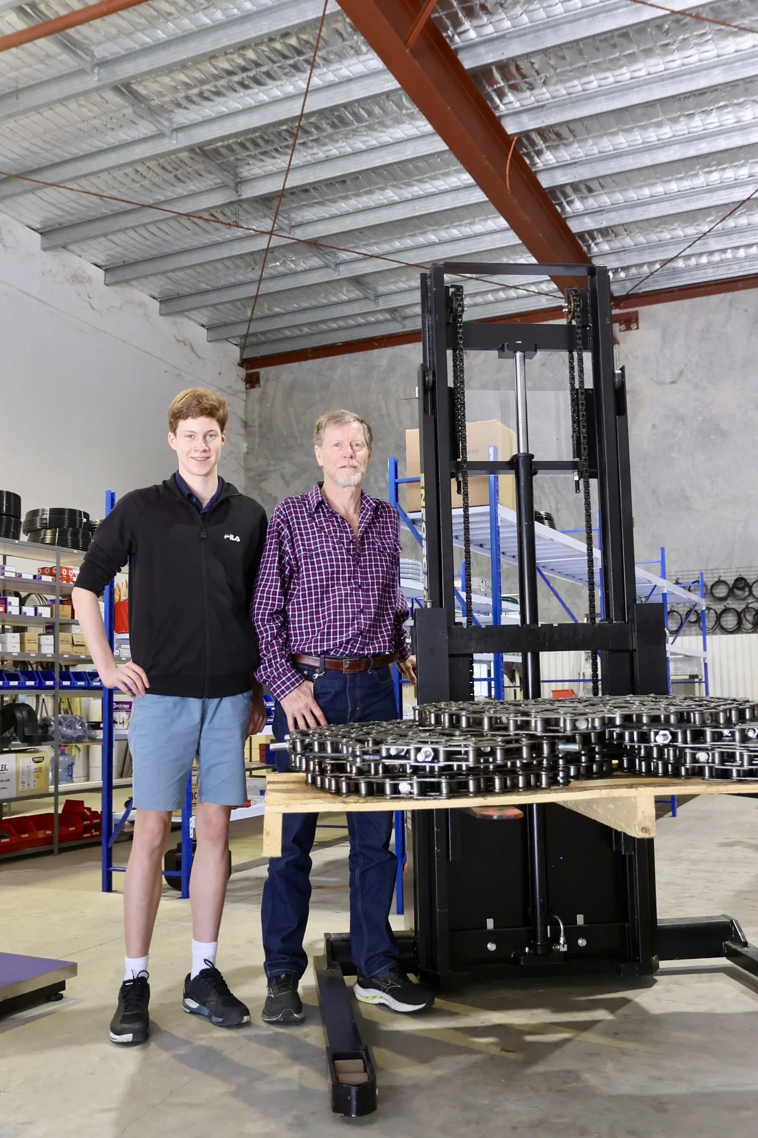 Two men standing in a warehouse next to a piece of industrial machinery, with shelves of supplies in the background.