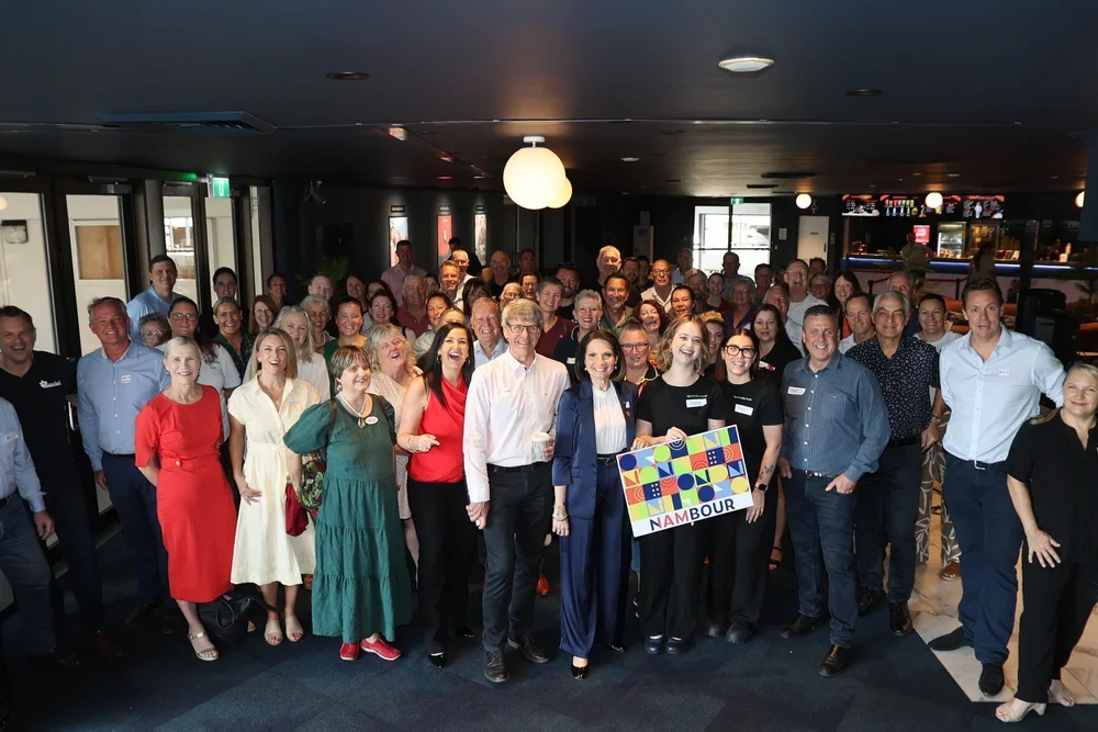 Group of people gathered indoors, some holding a colorful NAMBOUR sign, smiling for a group photo.