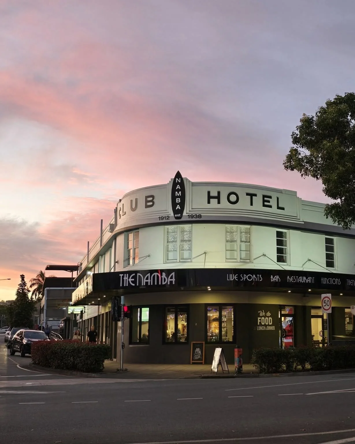 A street view of a building with a sign that reads "CLUB HOTEL" and a smaller sign for "THENAMBA" bar and restaurant, with sunset sky in the background.