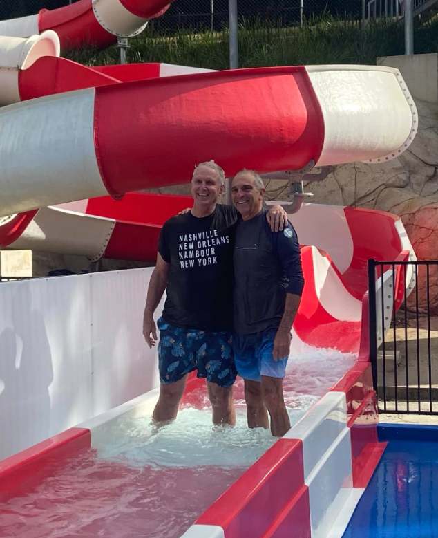 Two men standing in a shallow water pool at a water park, smiling, with a large red and white water slide in the background.