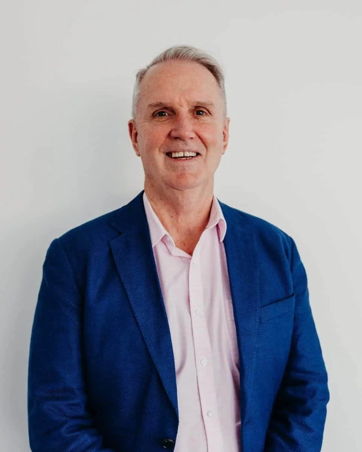 A smiling middle-aged man with light gray hair, wearing a dark blue blazer and a light pink collared shirt, standing against a plain white background.