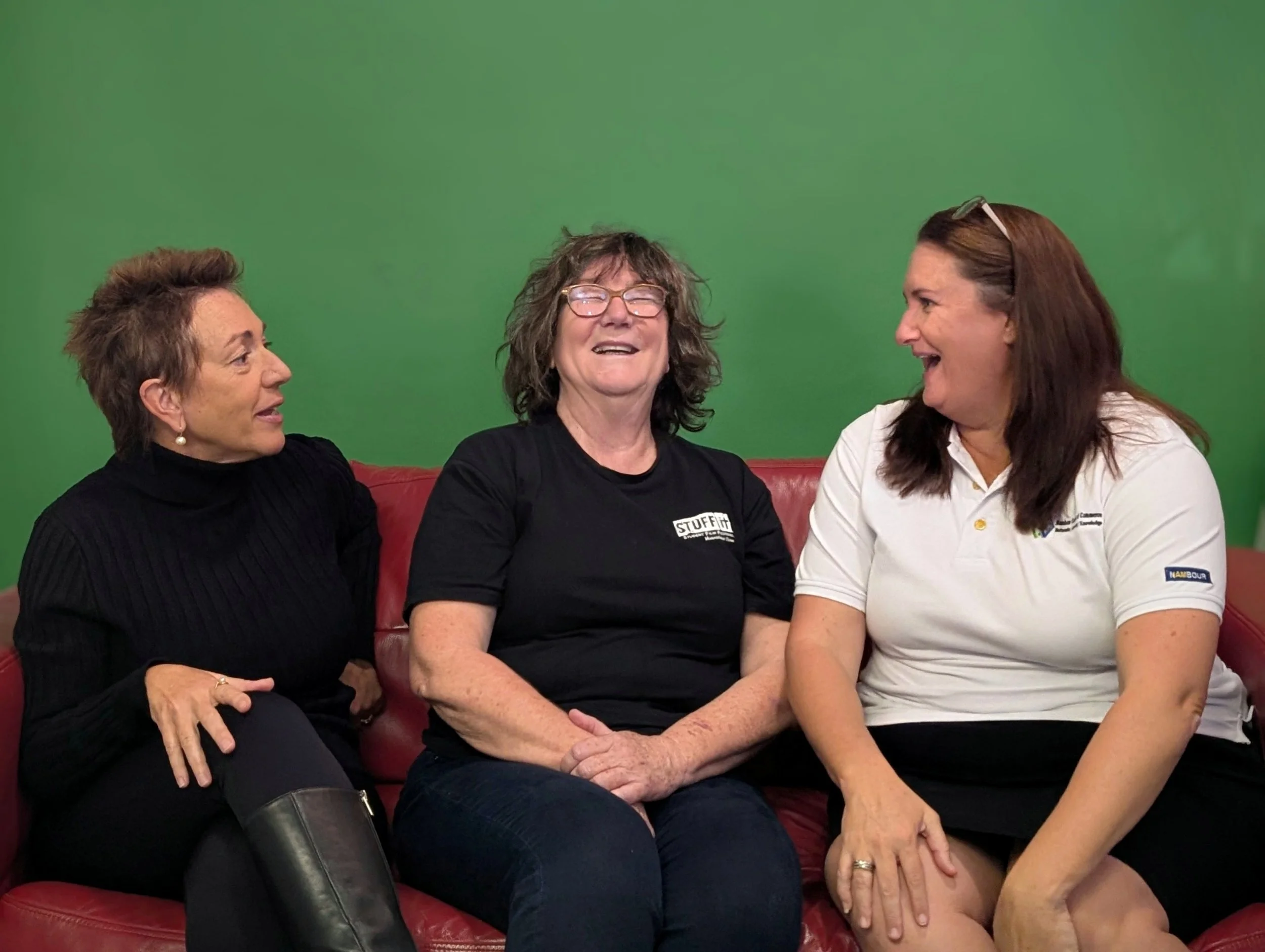 Three women sitting on a red couch in front of a green screen, smiling and talking to each other.
