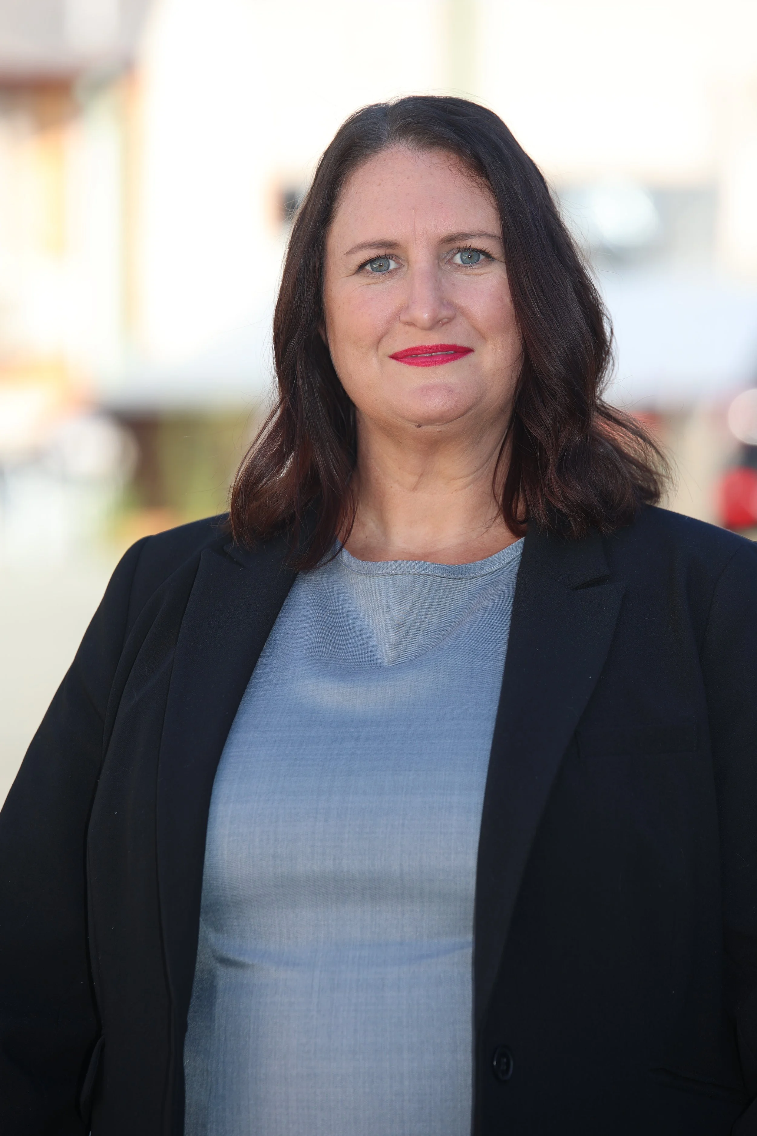 A woman with black hair and blue eyes wearing a dark blazer and light gray top, standing outdoors with a blurred background.