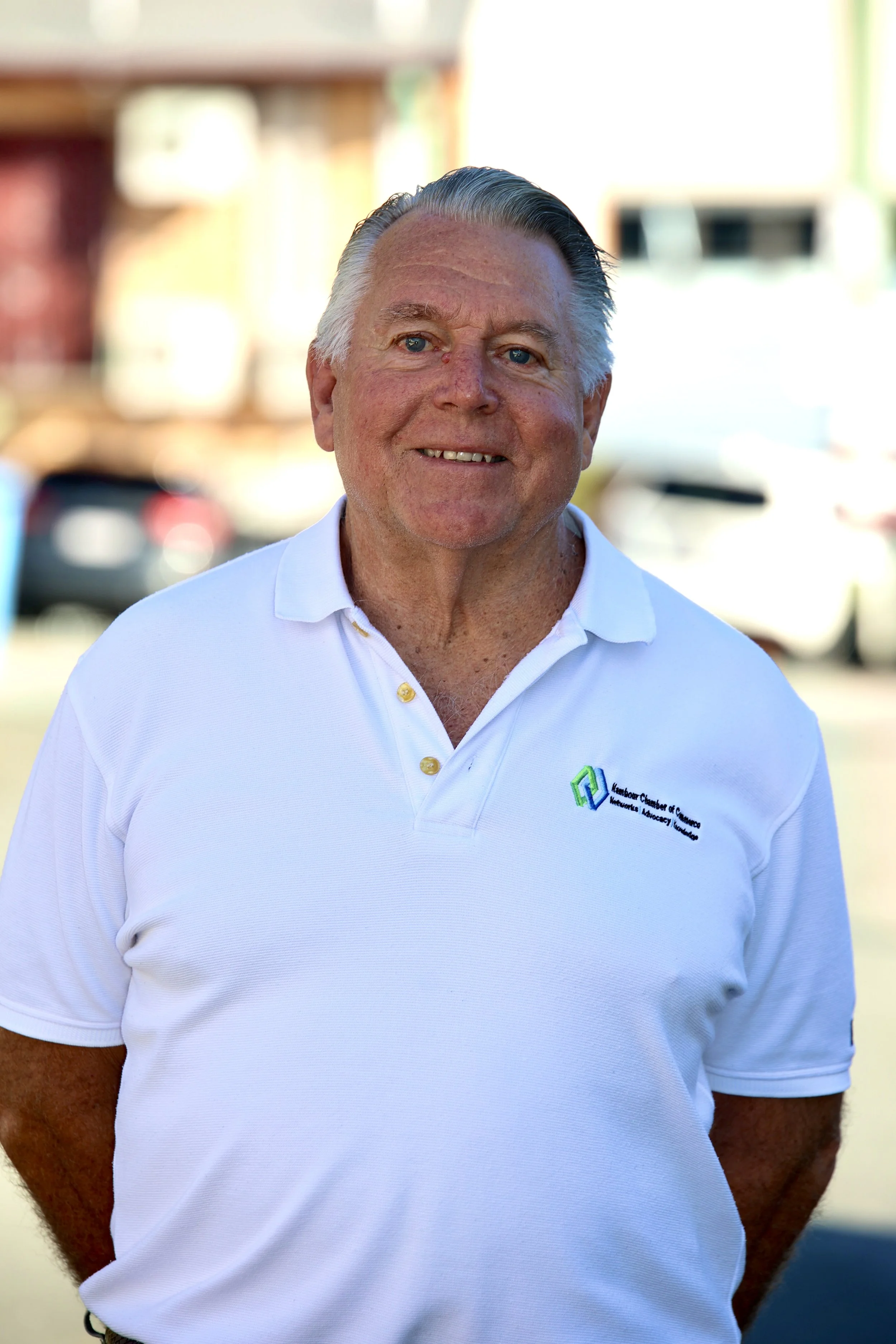 A middle-aged man with gray hair and blue eyes, wearing a white polo shirt with a logo, standing outdoors with a blurred background of buildings and cars.