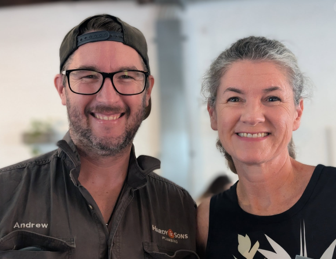 A smiling man wearing glasses, a dark jacket, and a cap, standing next to a smiling woman with gray hair in a black top.