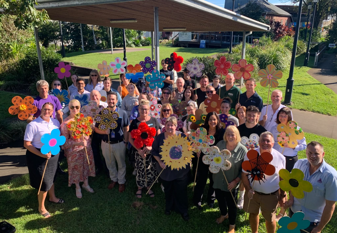 Group of people standing outdoors under a gazebo holding colorful flower-shaped signs and decorations, with a green lawn and pathway in the background on a sunny day.