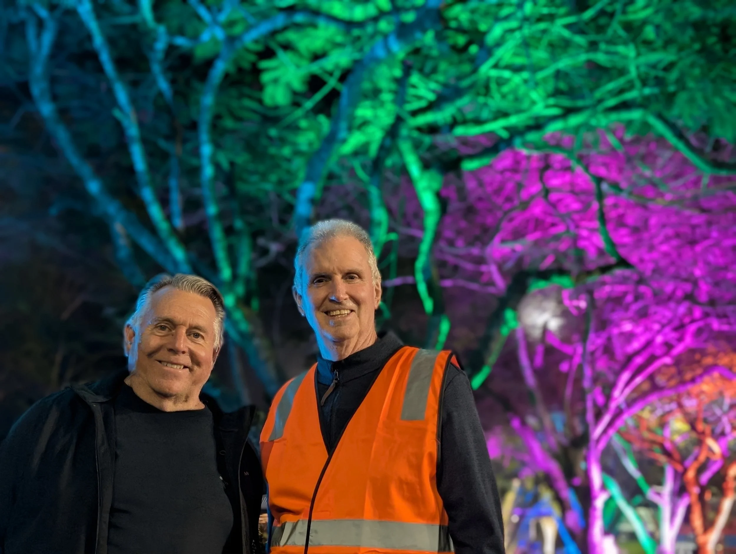 Three men standing in front of a colorful, illuminated tree with vibrant purple, green, and blue lights at night.