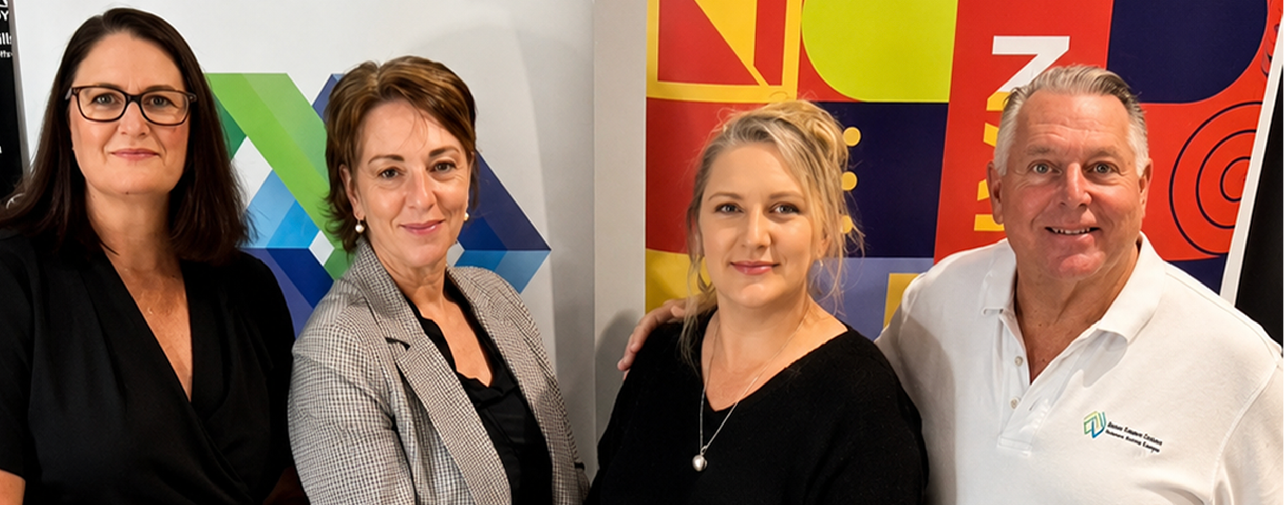 Four people standing together indoors in front of colorful geometric and abstract art. They are smiling and dressed in business casual attire.