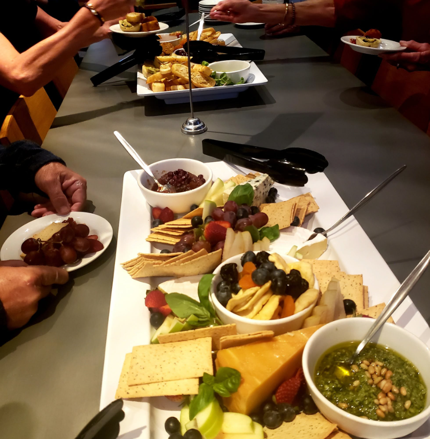 A group of people sharing a cheese platter with crackers, grapes, strawberries, blueberries, and small bowls of jelly and chutney at a dining table.