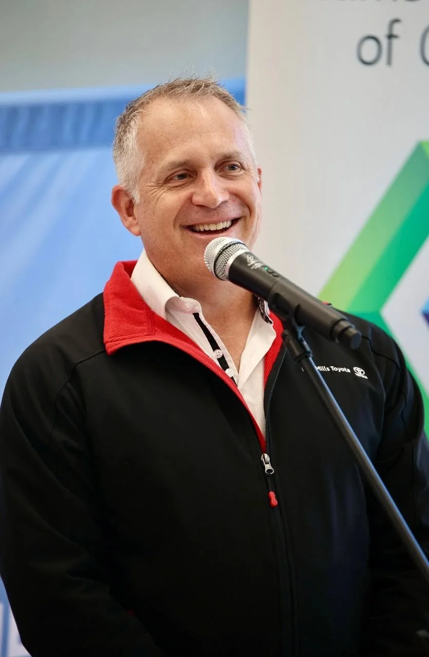 A man in a black and red jacket speaking into a microphone, smiling.