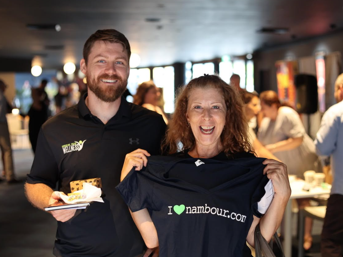A smiling woman holding a black "I love Nambour" shirt, standing next to a man in a black Wellness shirt, both at an indoor event with other people in the background.