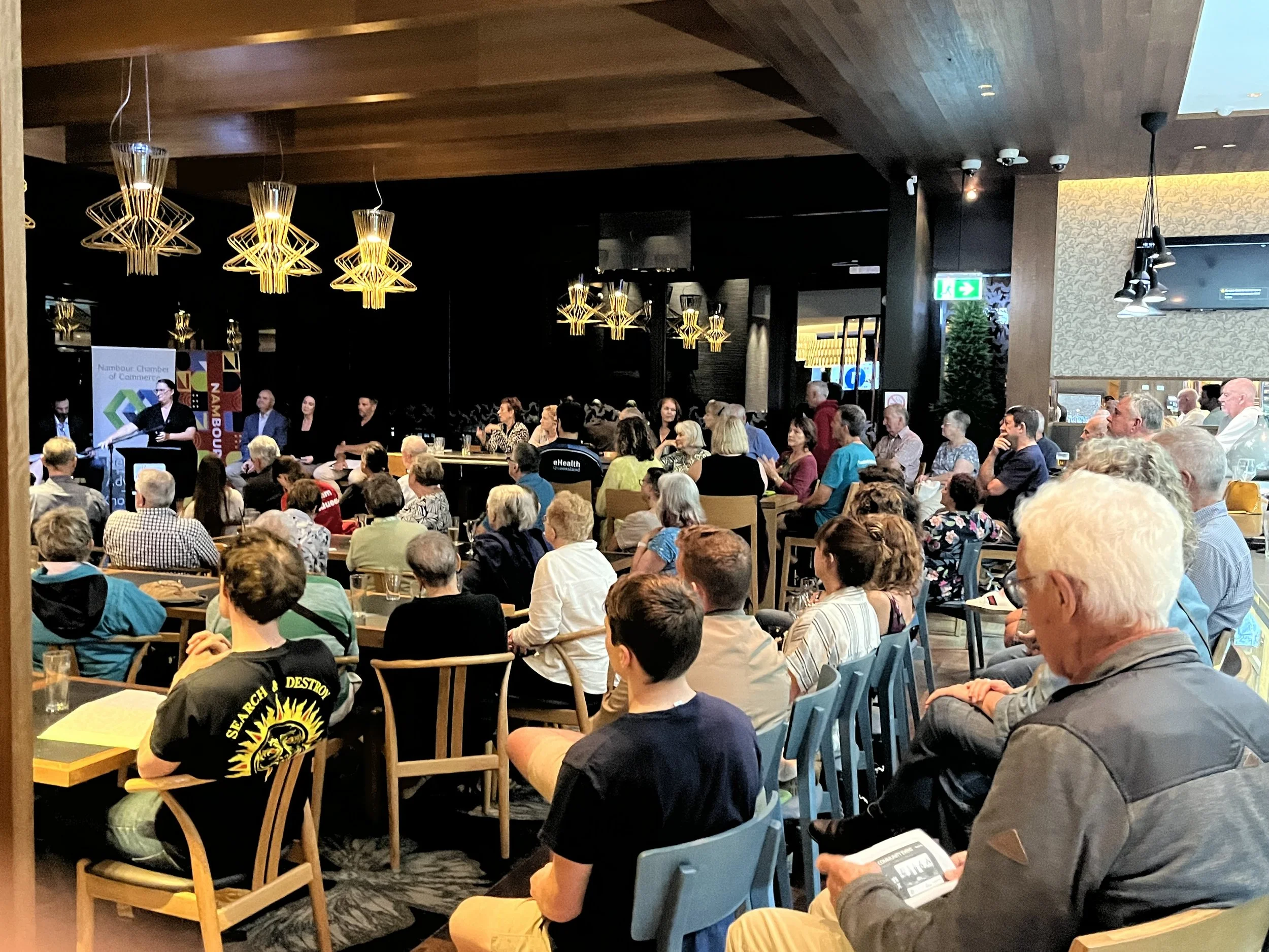 A large group of people sitting at tables in a conference or event room, facing a speaker or presentation at the front. The room features modern hanging light fixtures, wood-paneled ceilings, and a mix of wall decor and technology such as a TV screen