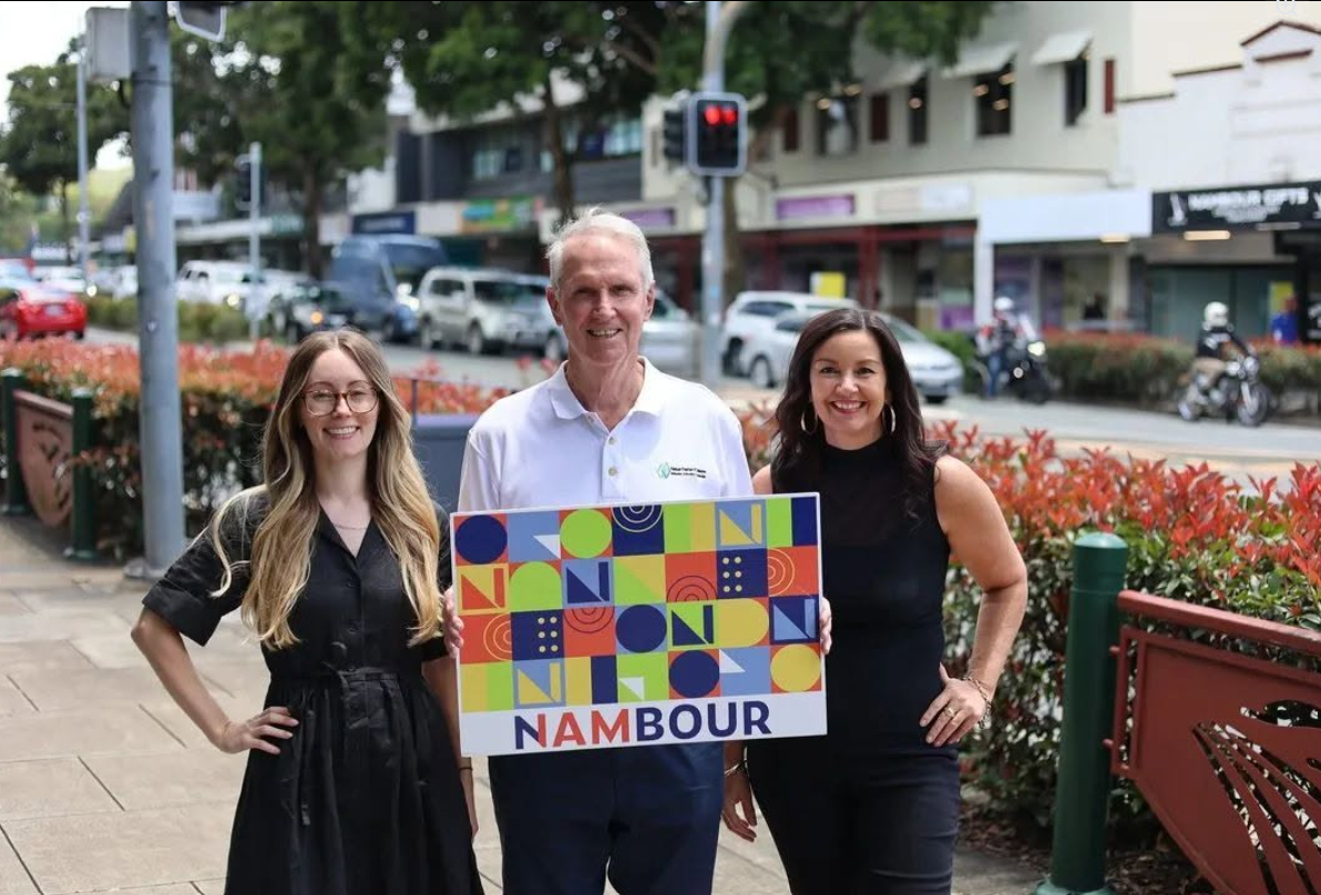 Three people standing outdoors on a city sidewalk, holding a colorful sign that says "NAMBOUR." The man in the middle wears a white polo shirt, and the two women on either side wear black outfits. Behind them are parked cars, bikes, and a row of shop