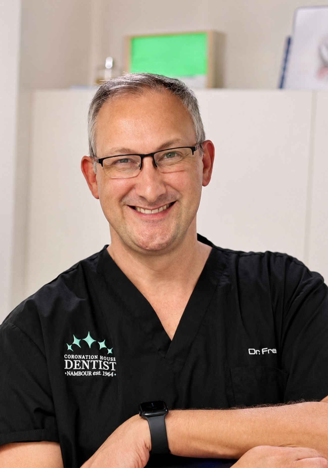 Smiling male dentist wearing glasses and black scrubs with logo, standing in dental office.