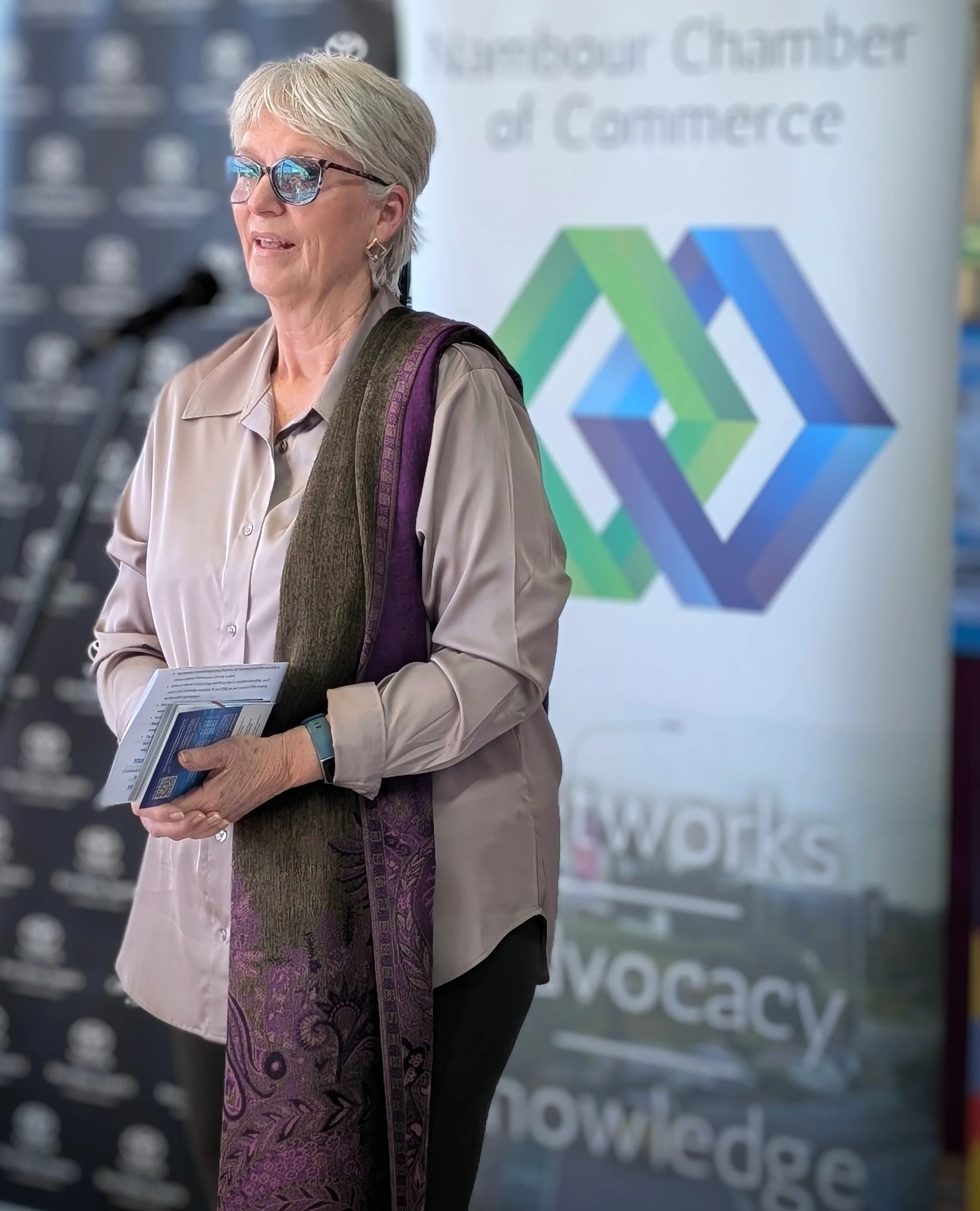 A woman with short gray hair wearing sunglasses, a beige blouse, and a purple patterned scarf, holding pamphlets, standing in front of a blurred sign that reads 'Nairobi Chamber of Commerce' and 'knowledge' and 'workplace'.