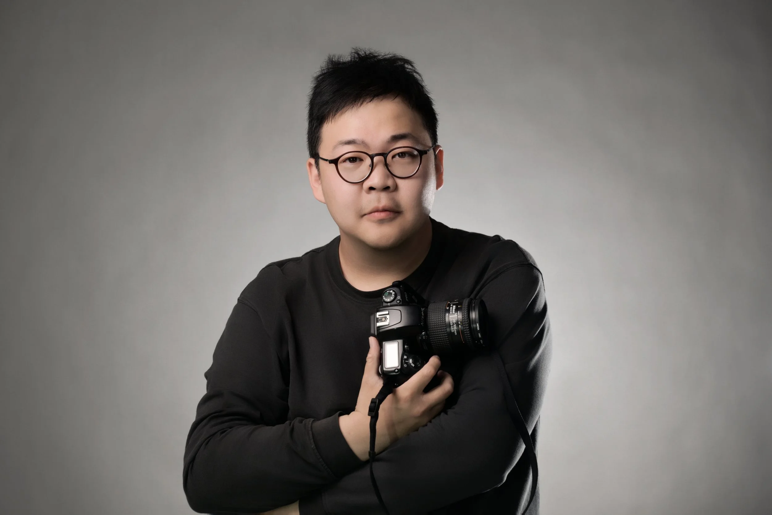 A young man with glasses and short black hair holding a camera in front of a plain grey background.