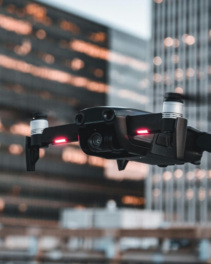 Black drone with camera flying in urban setting with tall buildings in the background.