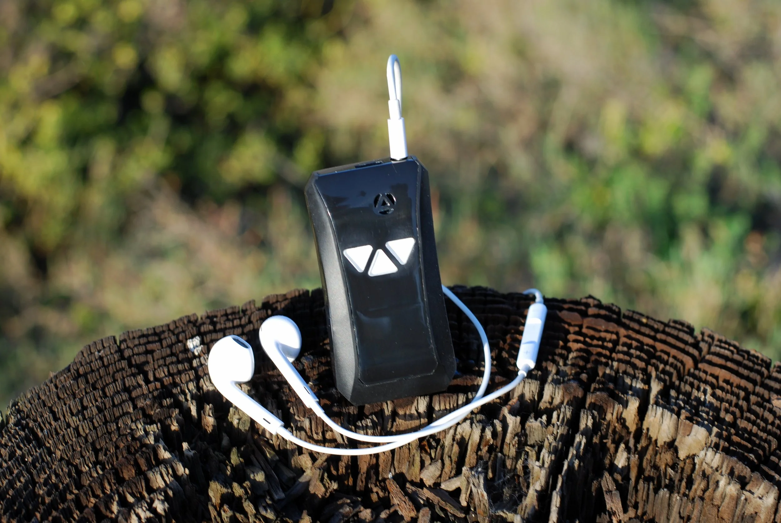A black remote control with white buttons and a headphone jack is placed on a cut tree trunk, with white earbuds and a charging cable attached, in an outdoor setting with green foliage in the background.