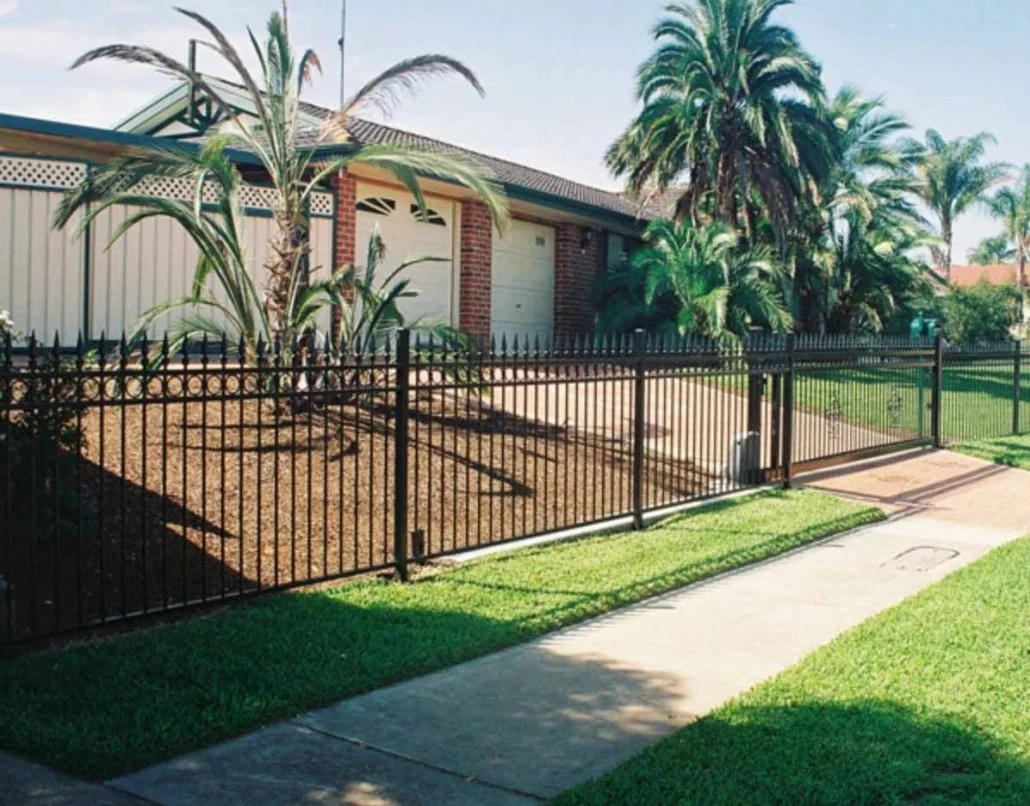 A suburban backyard with a black metal fence, a paved walkway, green grass, and various tropical plants including palm trees.