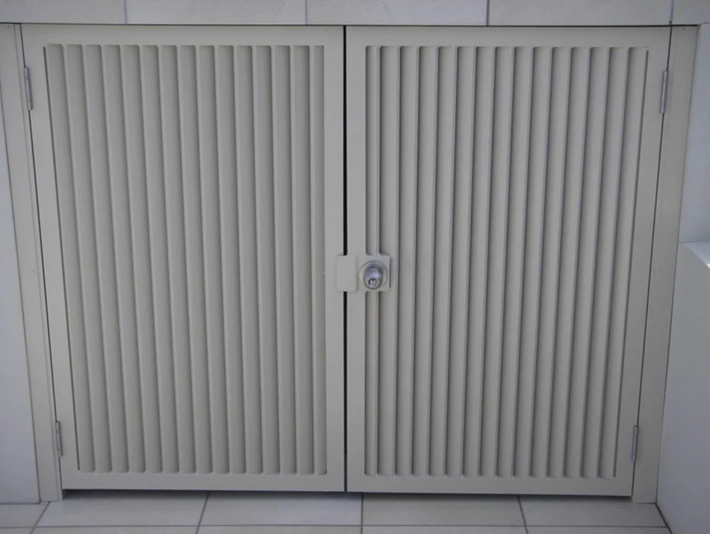 White metal cabinet with vertical slats, locked with a round lock in the center, mounted on a tiled wall.