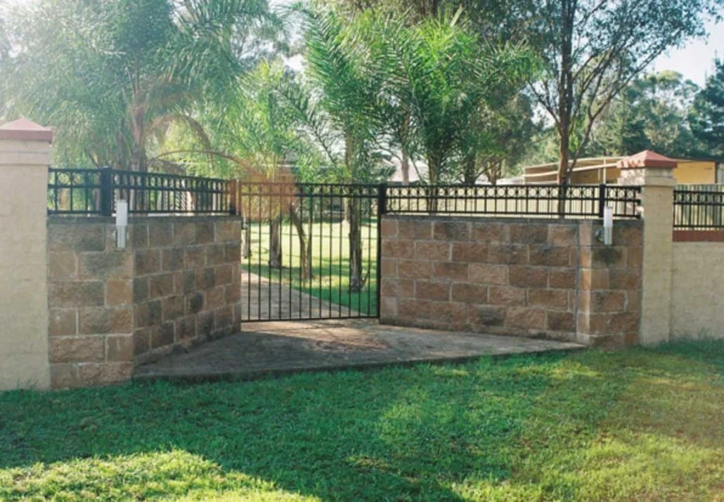 A gated entrance to a backyard with stone walls, a metal gate, and lush greenery including palm trees.