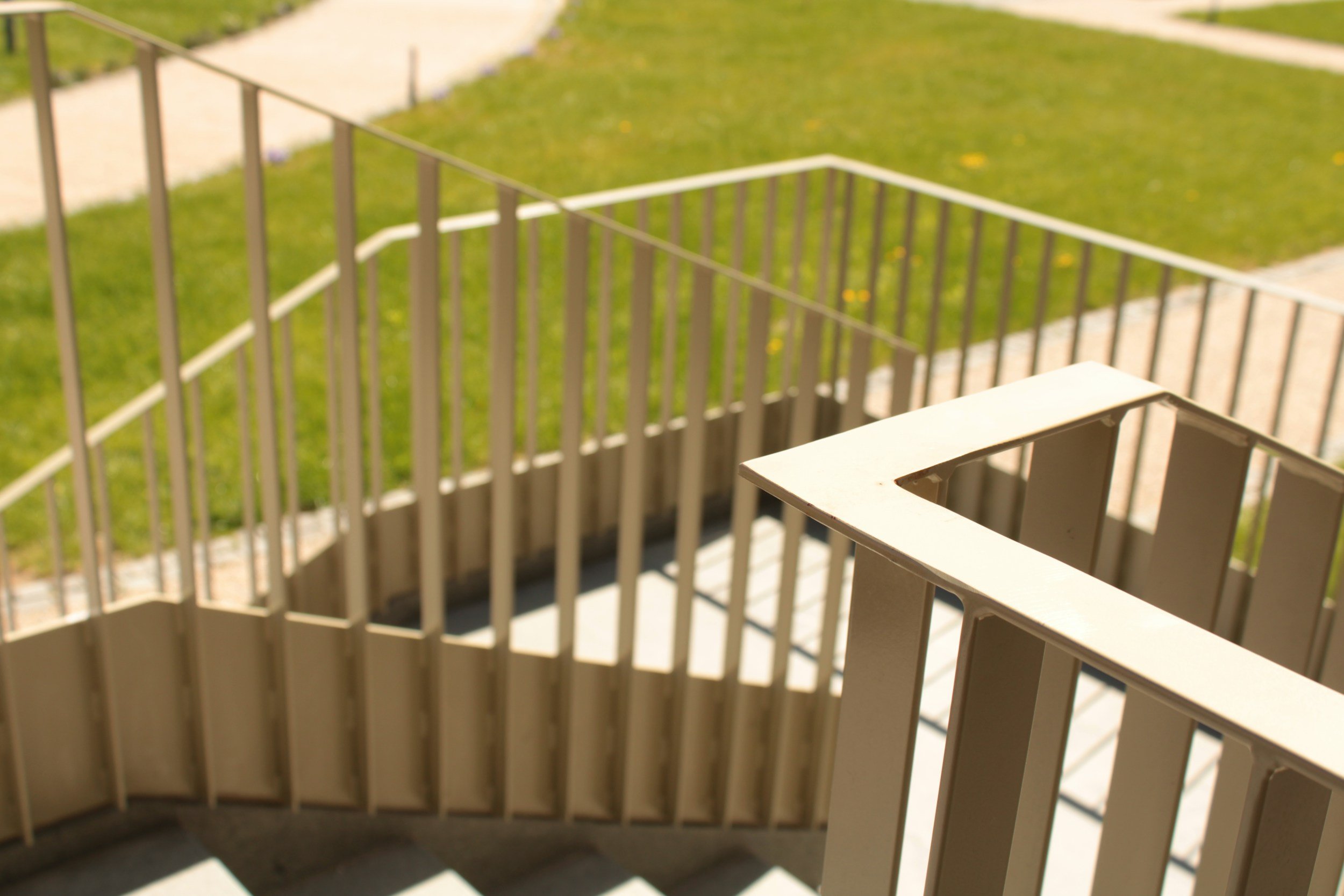 A metal staircase railing leading to a grassy lawn with yellow flowers, sidewalks, and a pathway in the background.