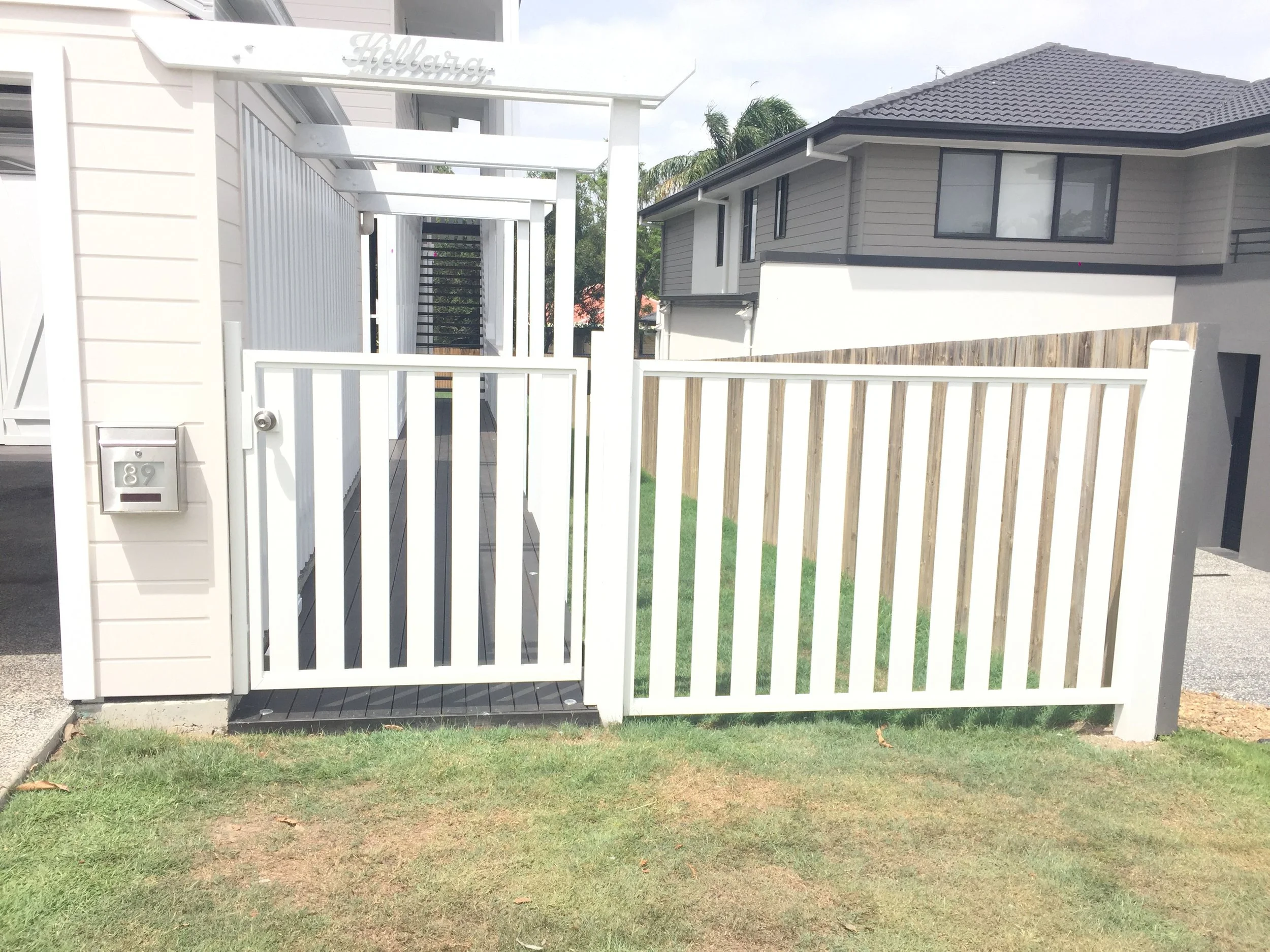 A white fence with a gate in the front yard of a house, a gray house with black windows in the background, a small patch of grass, and a wooden fence on the right side.