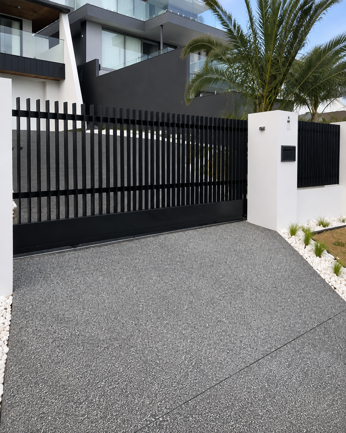 Modern black metal gate with white entrance pillar and mailbox, surrounded by white decorative rocks and a landscaped garden with a tall palm tree, in front of a contemporary residential building with glass balconies.