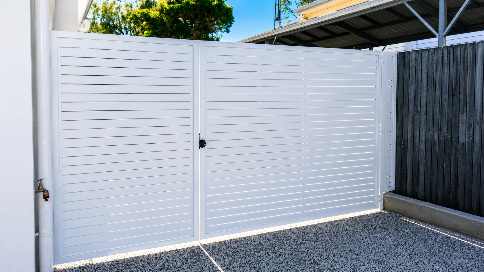 White metal security gate with horizontal slats, black door handle, and a black lock. Fencing includes a dark wooden panel on the right and a white wall on the left, with a gravel floor and partial view of a modern building with a roof overhang.