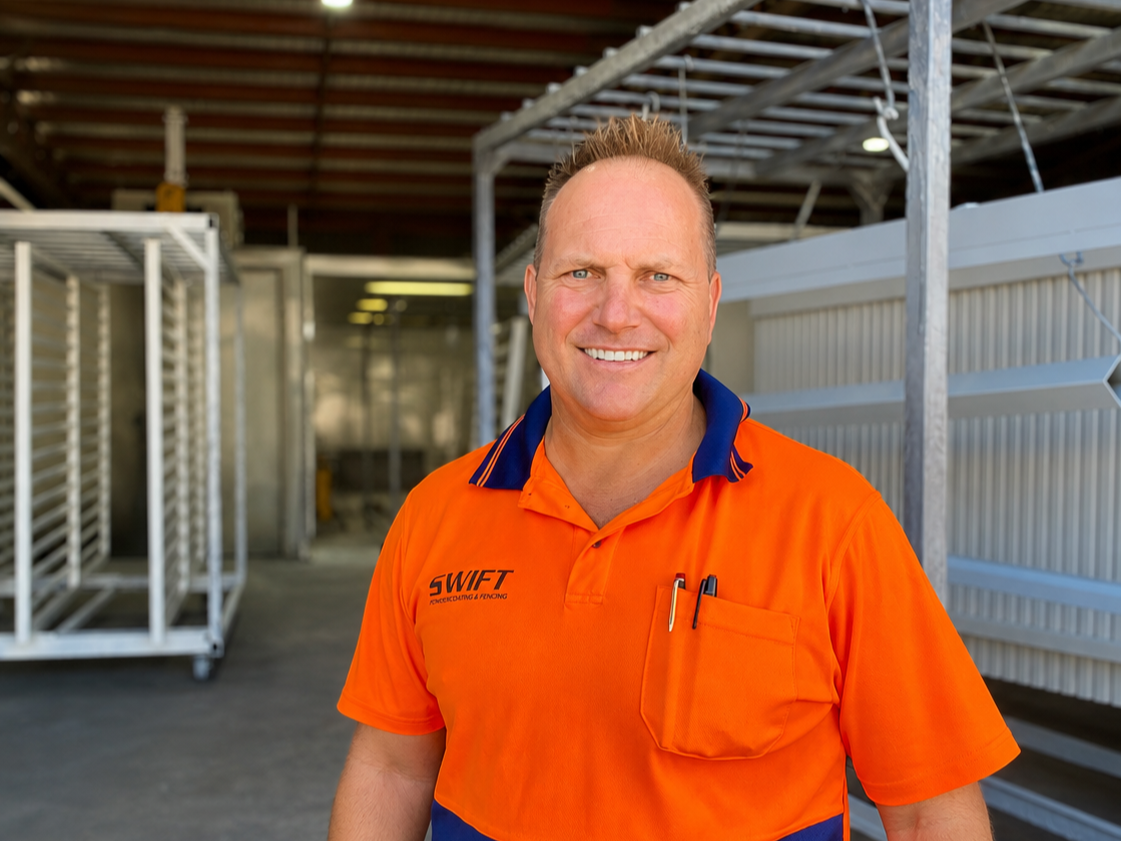 A man smiling at the camera in an industrial or warehouse setting, wearing an orange uniform shirt with the word 'SWIFT' on it.