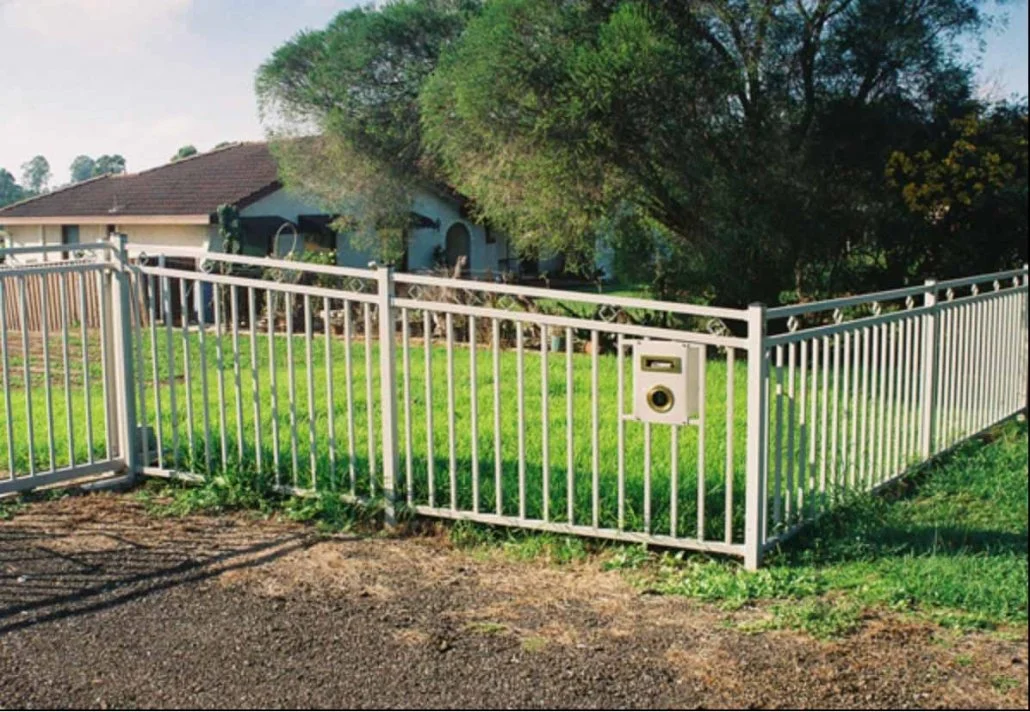 A white metal fence with a mailbox and mail slot in a grassy front yard of a house. The house has a brown roof and trees are in the background.