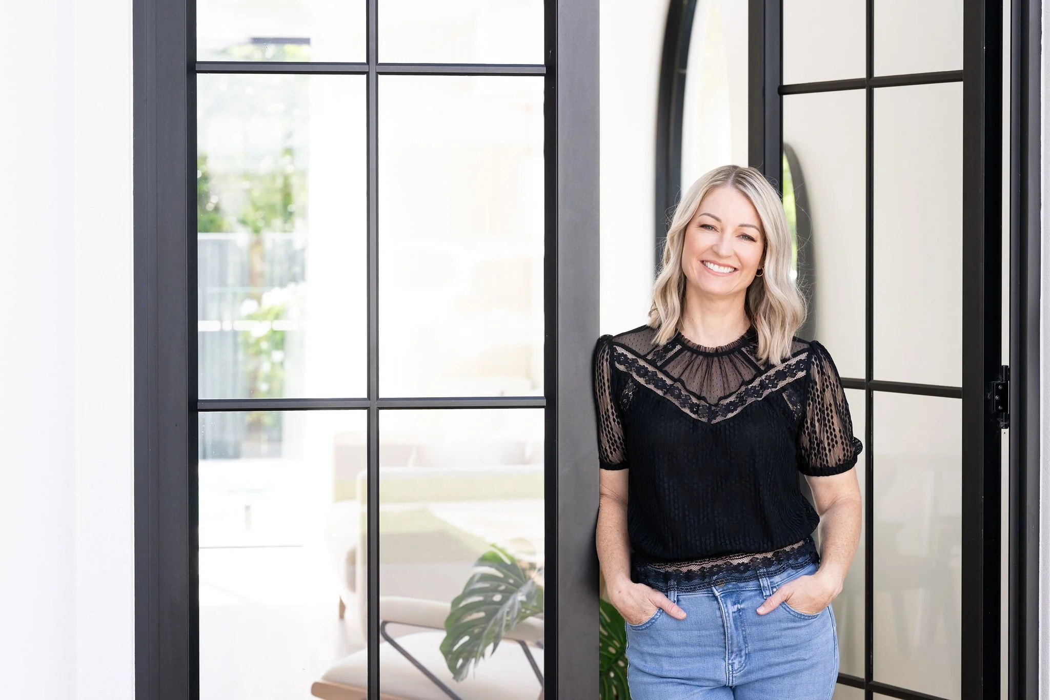 A woman with gray hair smiling and standing in front of glass doors with black frames, wearing a black lace top and blue jeans.