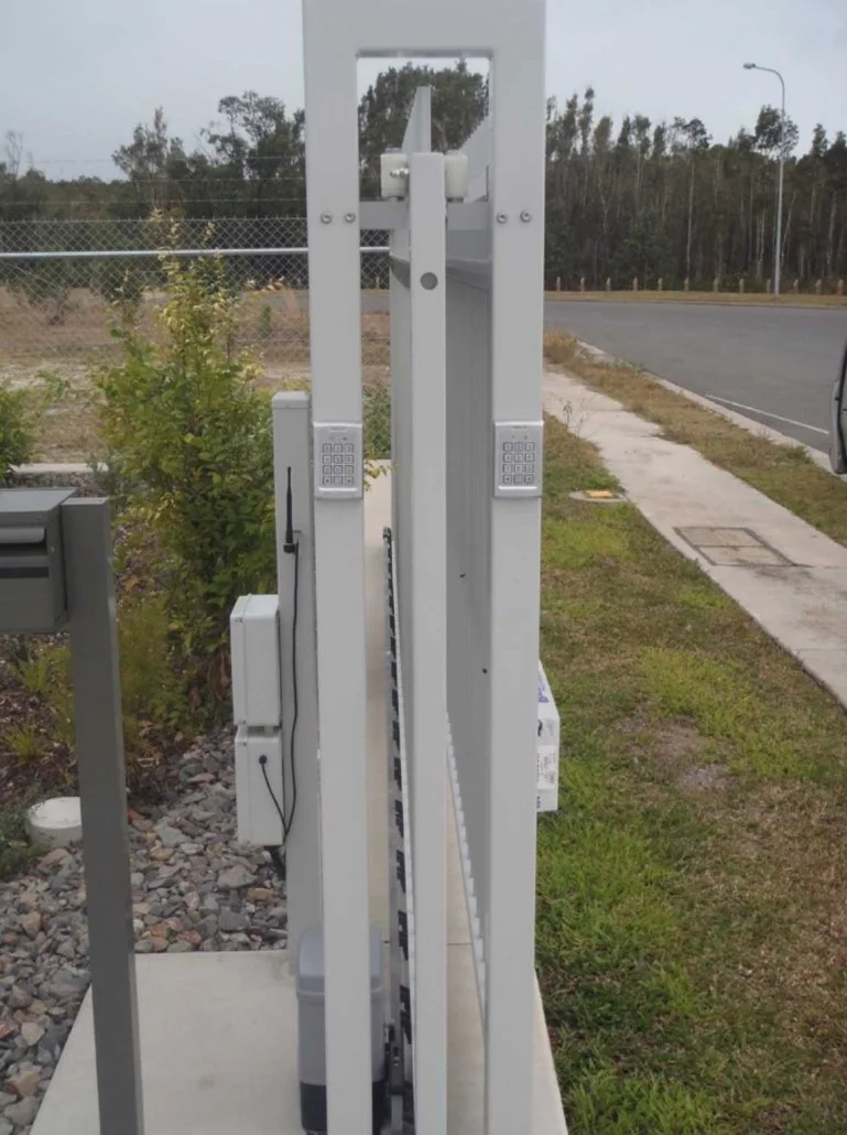 View of a white automation gate with keypad and motor, installed along a sidewalk next to a grassy area and street, with trees and a fence in the background.
